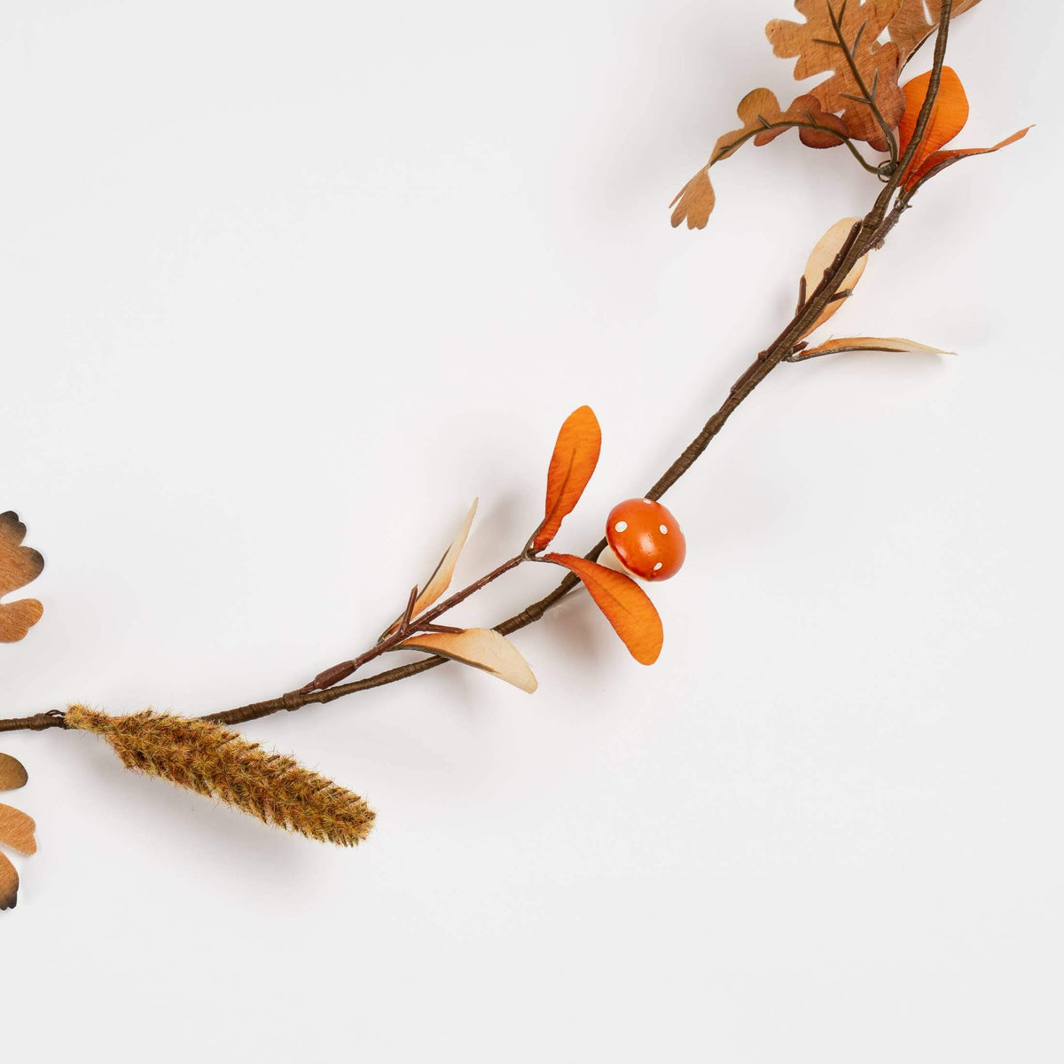 An autumn garland with orange berries, brown and orange leaves, and a brown faux-wood plume, laid diagonally on a plain white background.