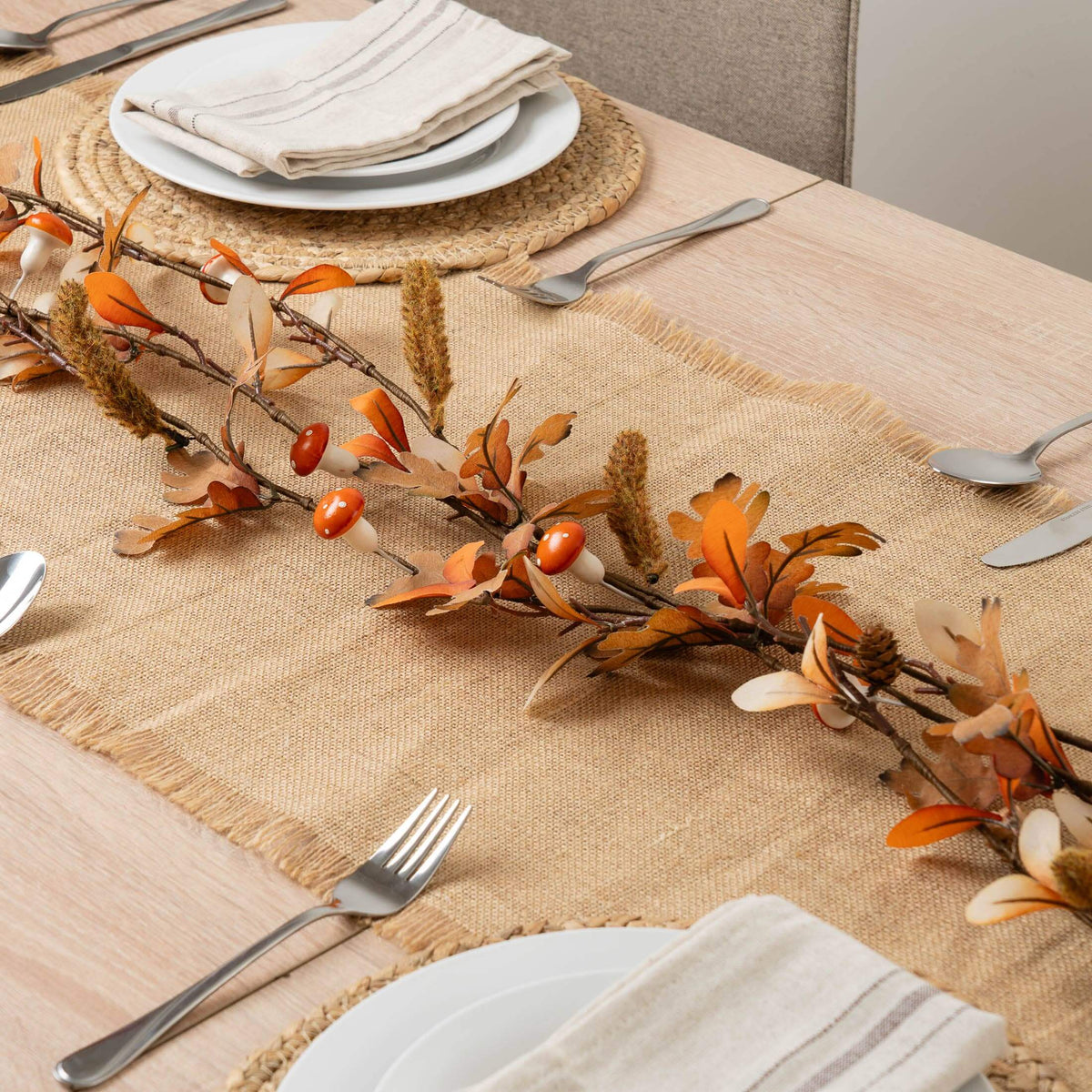 An autumn-themed table runner made of natural burlap is styled on a light wood dining table with white plates, folded cream linen napkins, silver cutlery, and a decorative garland featuring orange leaves, brown branches, and small red berries.