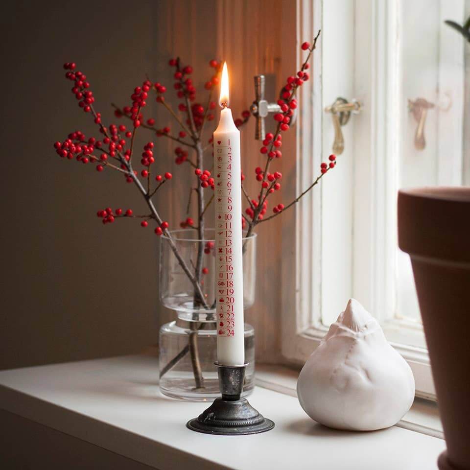 A white taper candle with red printed text stands lit in a black metal holder on a windowsill, surrounded by red berries in a glass vase and a white ceramic bird figurine.