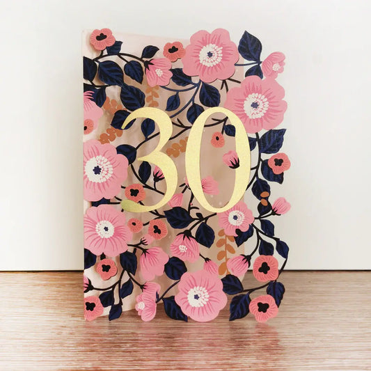 A floral birthday card with '30' printed in gold, surrounded by pink flowers and green leaves, with a brown kraft envelope.