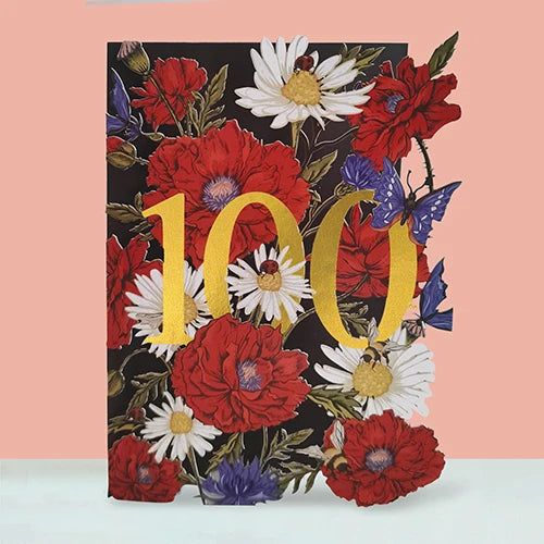 A square decorative card featuring a floral design with large red and white flowers, blue butterflies, and bold gold '100' lettering on a black background, set against a peach wall and white surface.