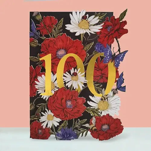 A square decorative card featuring a floral design with large red and white flowers, blue butterflies, and bold gold '100' lettering on a black background, set against a peach wall and white surface.