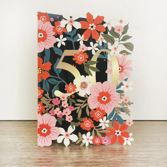 A colorful floral 50th birthday card with gold foil embellishments, standing upright on a wooden surface with a brown kraft envelope behind it.