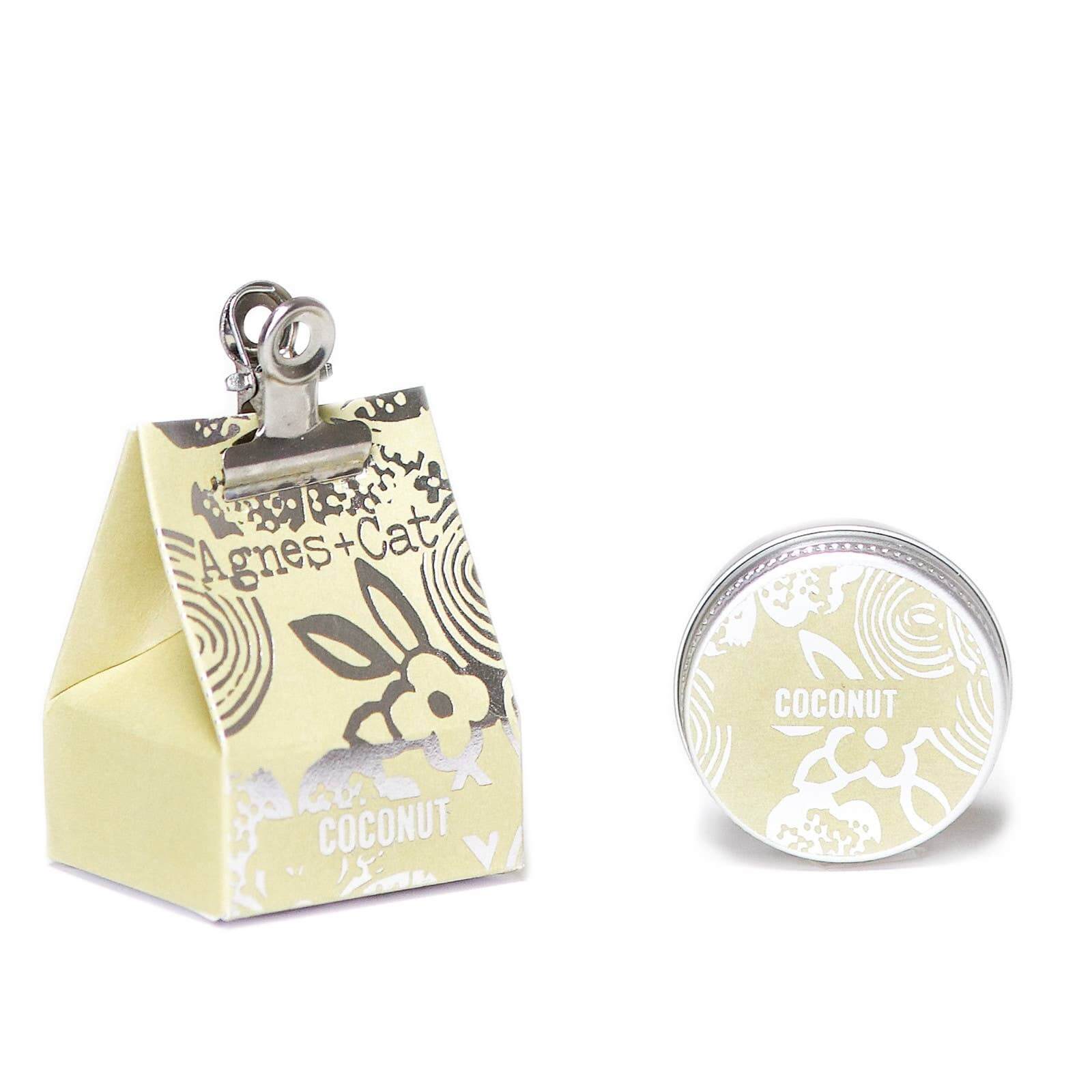 A small tin container of Agnes and Cat Coconut lip balm with a decorative design, showing the coconut fragrance and ingredients like shea butter and mango oil. It is accompanied by a box in the same design.