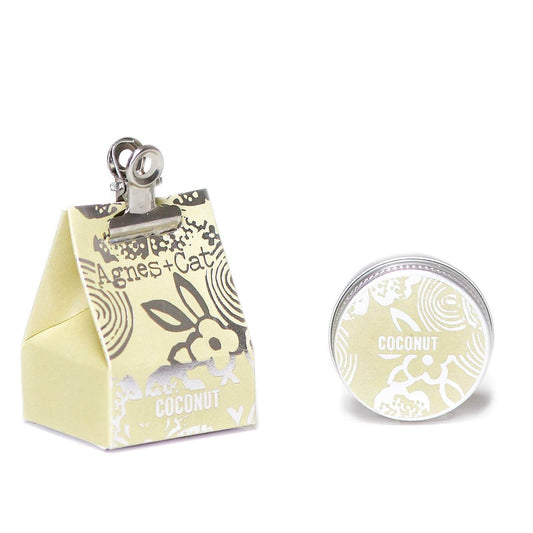 A small tin container of Agnes and Cat Coconut lip balm with a decorative design, showing the coconut fragrance and ingredients like shea butter and mango oil. It is accompanied by a box in the same design.