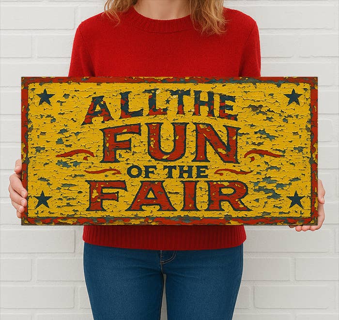 Person holding a vintage-style sign with 'All the Fun of the Fair' text against a white brick wall.