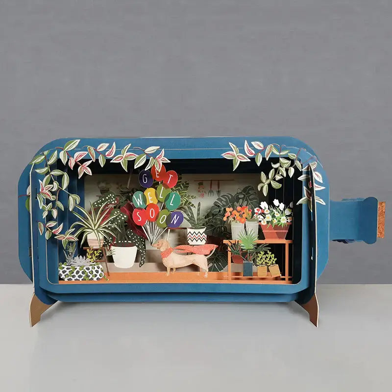 A 3D pop-up greeting card depicting a scene with plants and the message 'SOON GET WELL' in bold letters, set against a blue background.