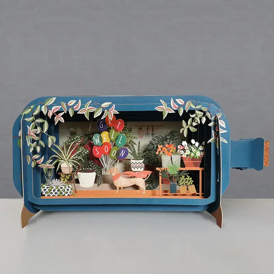 A 3D pop-up greeting card depicting a scene with plants and the message 'SOON GET WELL' in bold letters, set against a blue background.