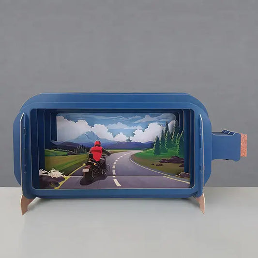 A pop-up greeting card depicting a motorbike on a road, with a person riding it towards mountains in the background. The card is blue with a die-cut paper technique.