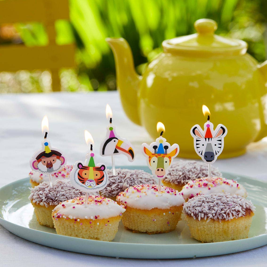 Cupcakes with animal-themed candles on a plate in front of a yellow teapot.