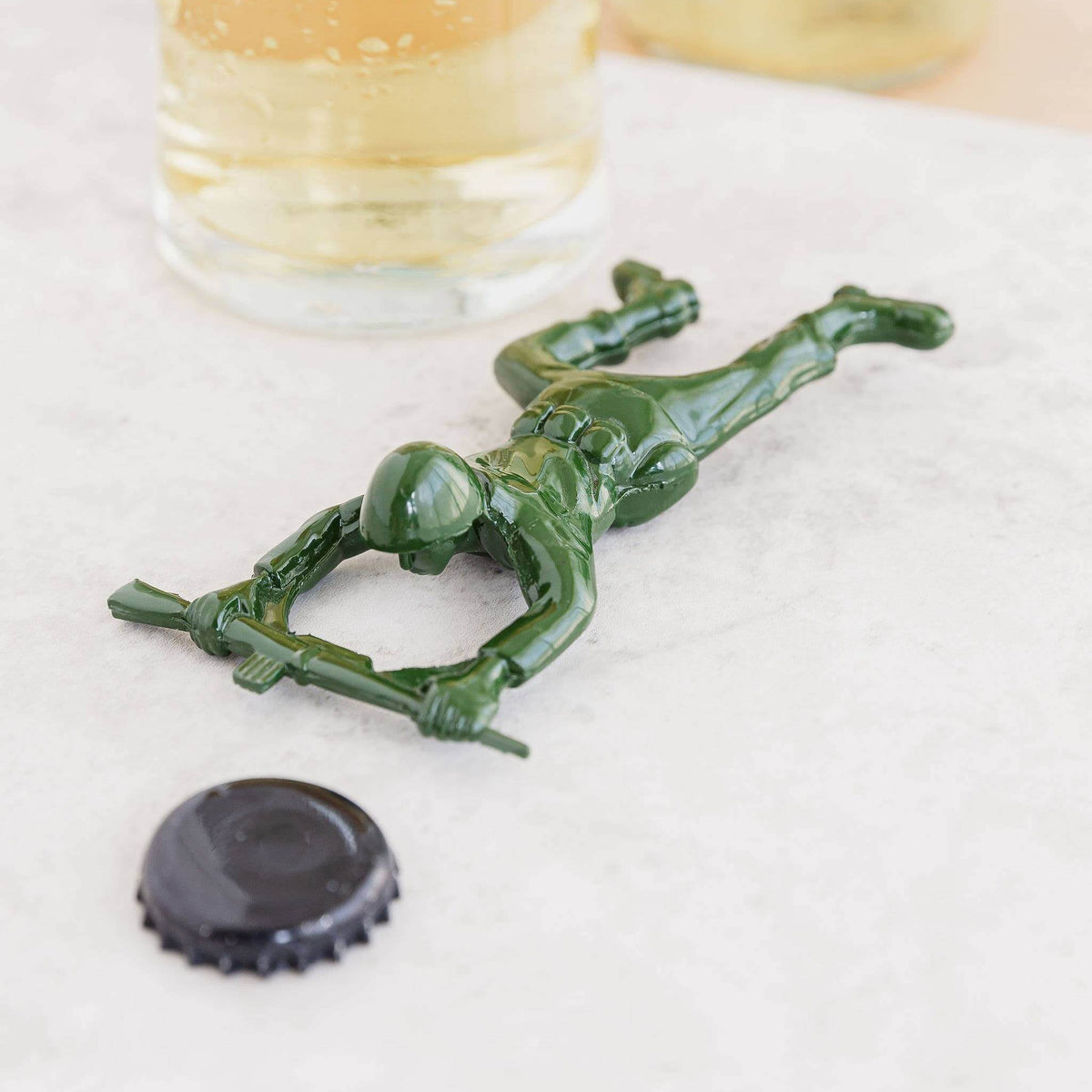 Green toy soldier bottle opener on a white surface with a glass of beer and bottle cap nearby.
