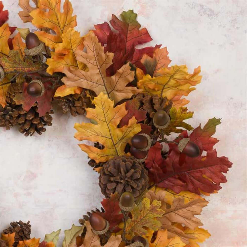 An autumn-themed decorative wreath made of faux oak leaves in shades of yellow, orange, red, and brown, accented with pinecones and acorns, photographed on a light textured background.