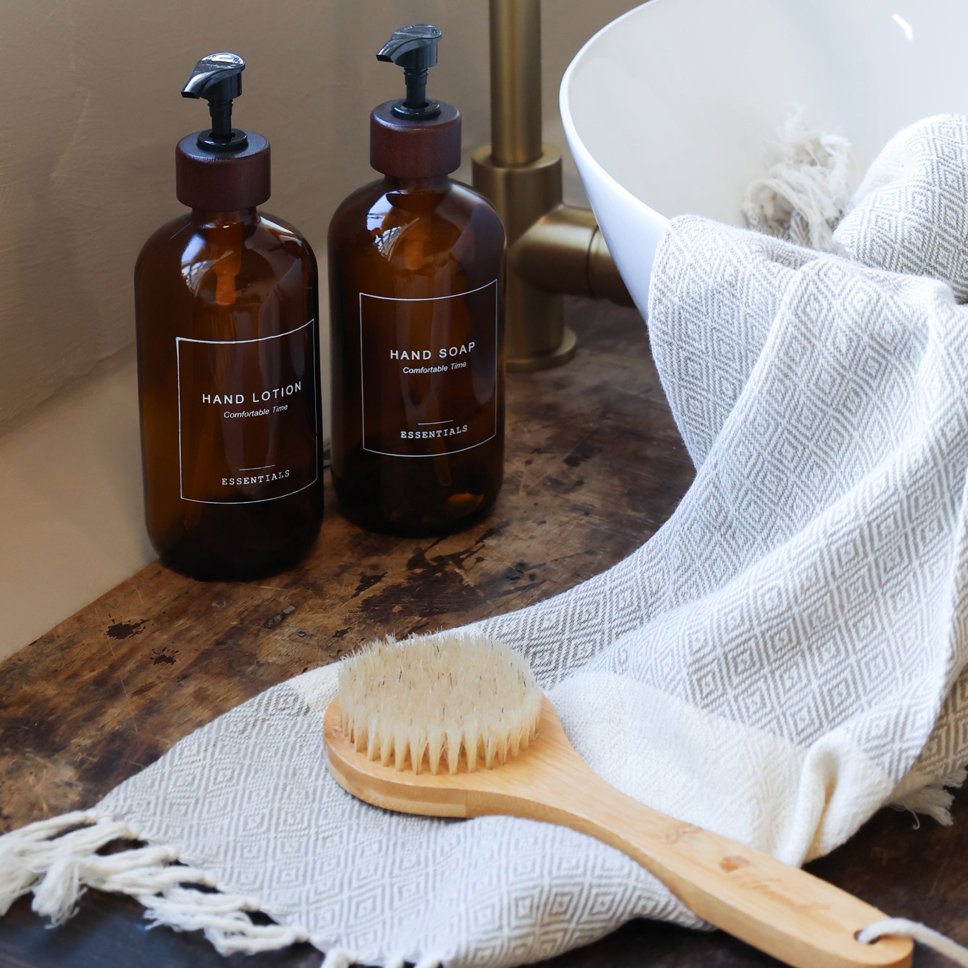 Two brown bottles labeled 'Hand Lotion' and 'Hand Soap' on a wooden surface with a white towel and wooden brush.
