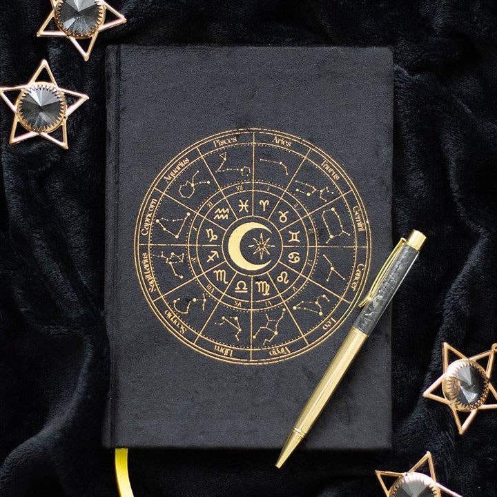 Black notebook with gold astrological design, gold pen, and decorative stars on a dark background