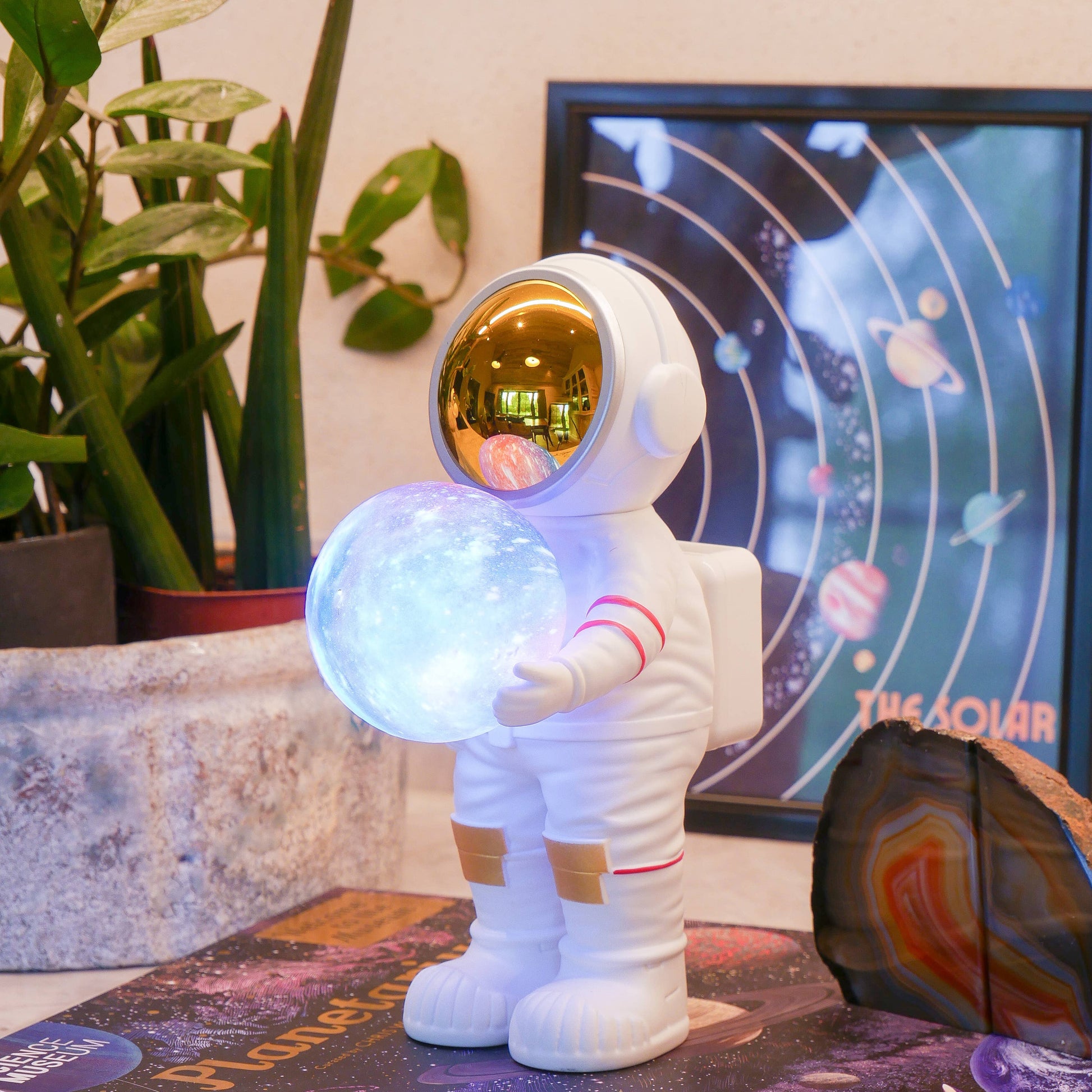 Spaceman figurine holding a glowing sphere on top of a 'Planetarium' book with a plant and framed picture in the background.