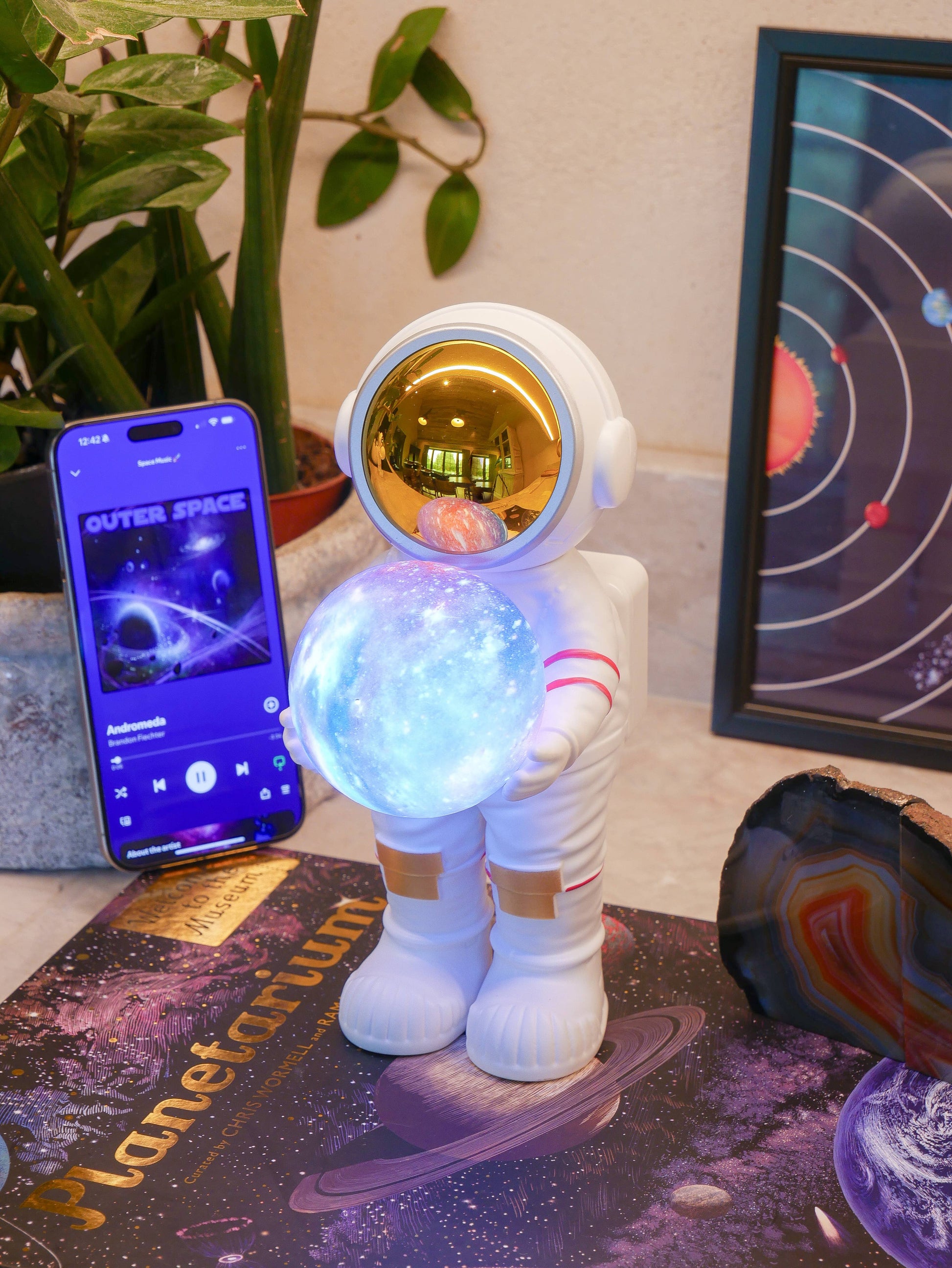 Toy robot bluetooth speaker with glowing blue ball on a table next to a smartphone displaying an outer space app.