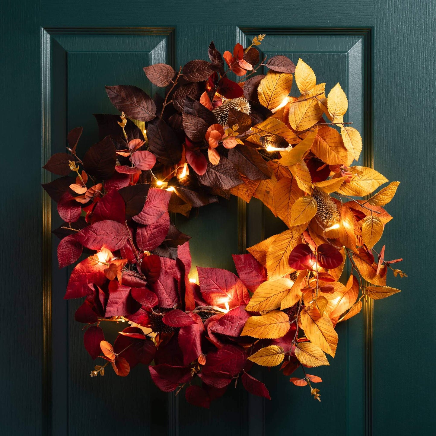 An autumn-themed artificial wreath with glowing warm white string lights, featuring a split design of deep burgundy and golden yellow leaves, hanging on a dark teal paneled door.