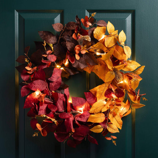 An autumn-themed artificial wreath with glowing warm white string lights, featuring a split design of deep burgundy and golden yellow leaves, hanging on a dark teal paneled door.