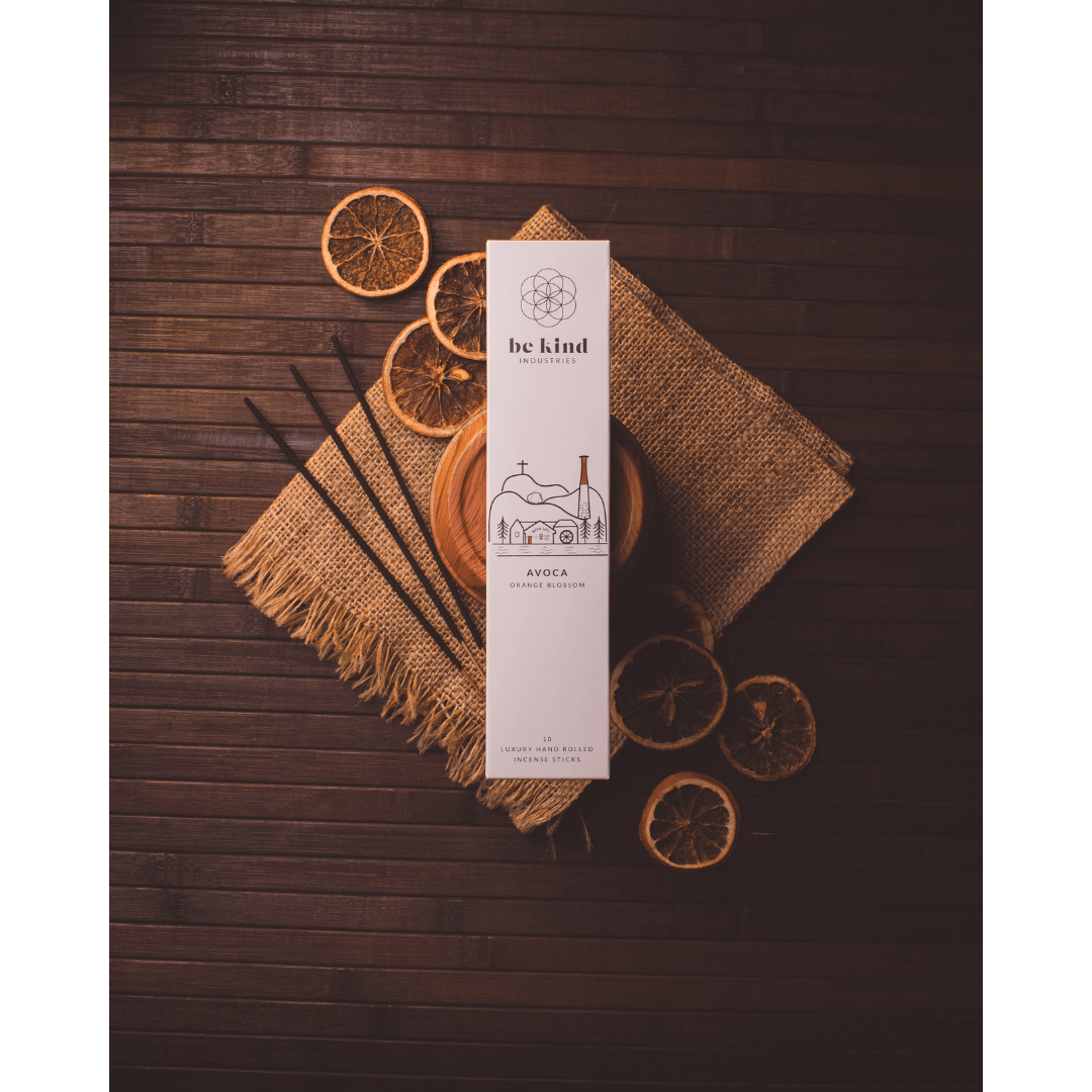 A package of Be Kind brand incense sticks, labeled 'Avoca- Orange Blossom' sitting on a wood surface with a jute tablemat and dried orange slices.