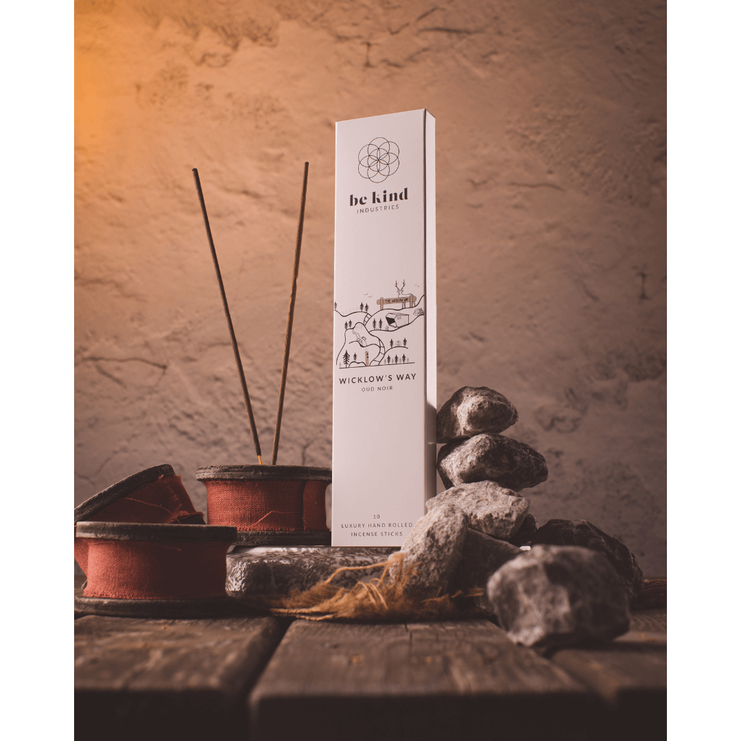 A package of Be Kind industries Wicklow's Way incense sticks. The packaging is primarily white with a graphic design featuring a house and a tree, and text detailing the product name and length. Situated on a rustic shelf with rocks and incense sticks