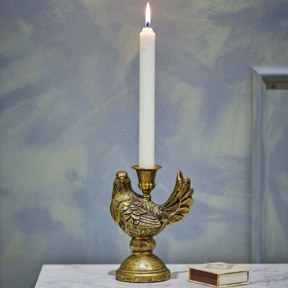 An ornate gold-toned bird-shaped candle holder holding a lit white taper candle, set on a marble surface against a textured blue-gray wall.