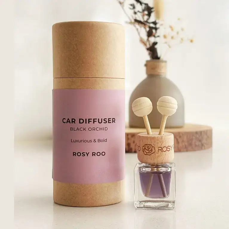 A small glass reed diffuser bottle with a light wood cap and two wooden diffuser sticks, placed on a white surface in front of a ceramic vase with dried stems on a round wood slice.