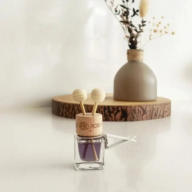 A small reed diffuser bottle with a cork top printed with the brand name 'Blossom Rose' and two wooden diffuser sticks, placed on a white surface in front of a vase with dried stems on a wood slice.