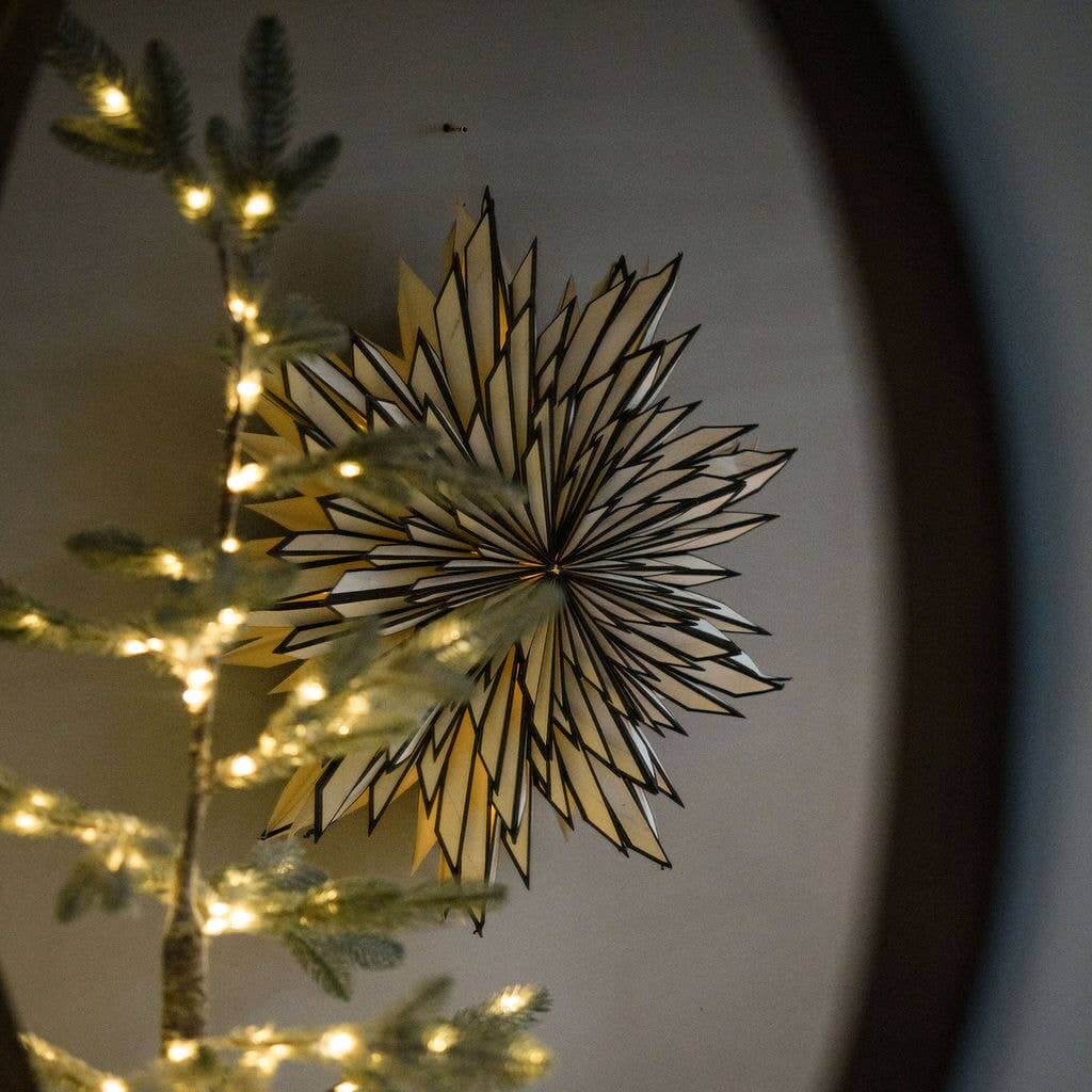 A large, circular, black and white paper wall decoration with a floral pattern, hanging on a wall with a Christmas tree and lights visible in the background.