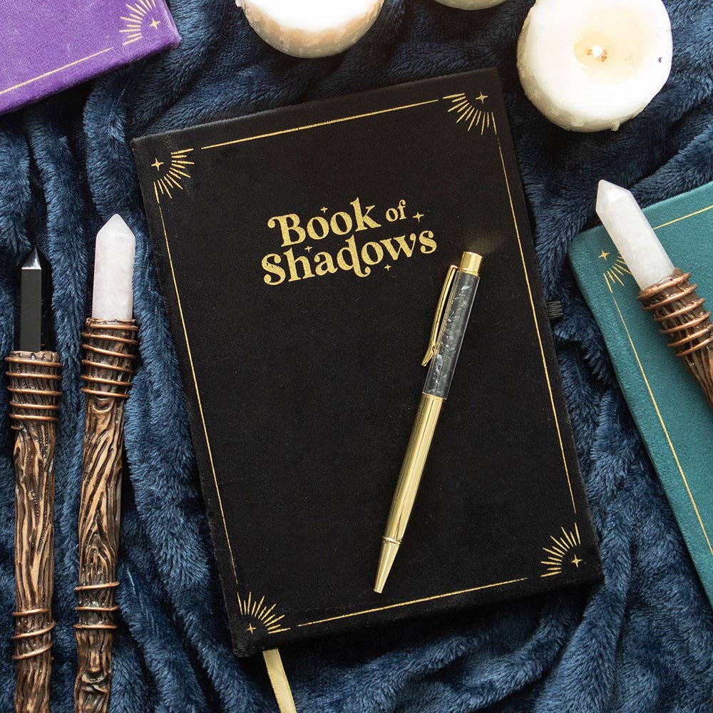 Black notebook titled 'Book of Shadows' with gold text, surrounded by candles and decorative items on a dark blue fabric background.