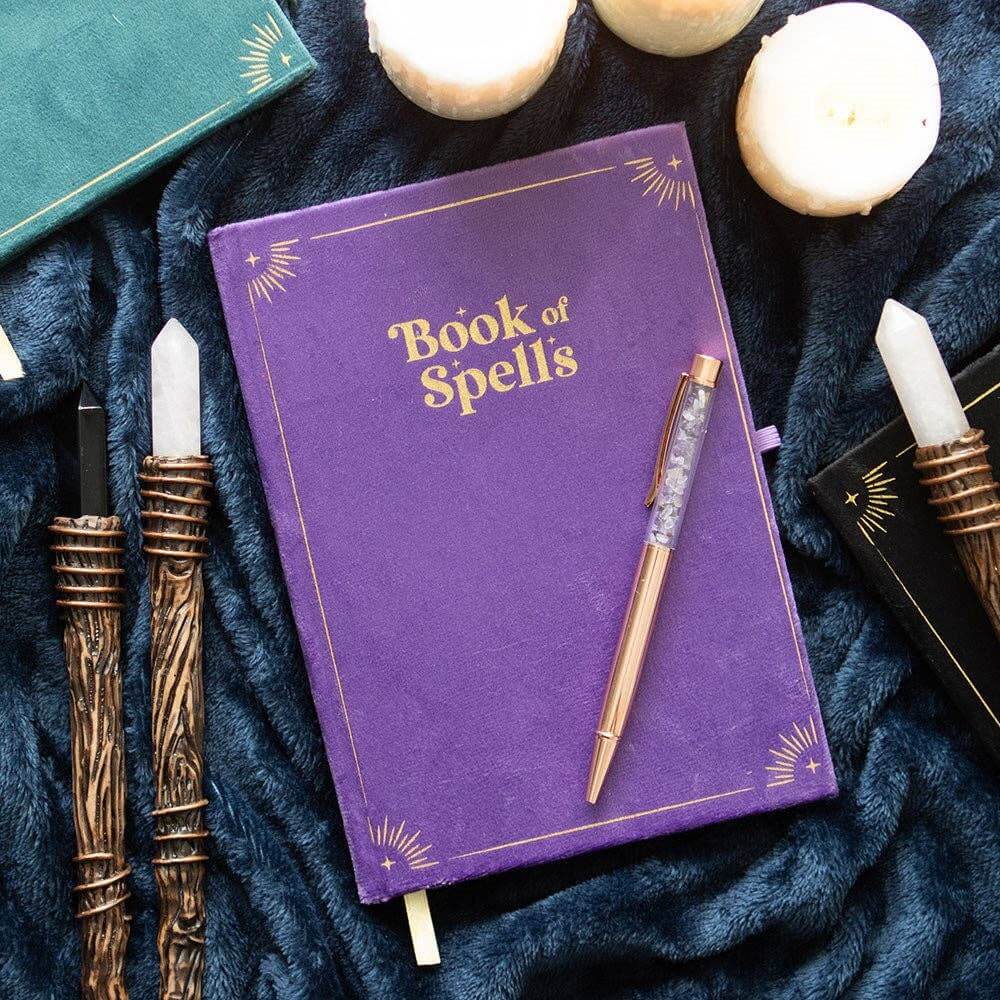 Purple notebook titled 'Book of Spells' with a pen on a dark blue fabric background with candles and wands.