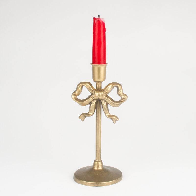 A vintage-style gold brass candlestick holder with a decorative bow motif on the stem, holding a single red taper candle, set against a plain white background.