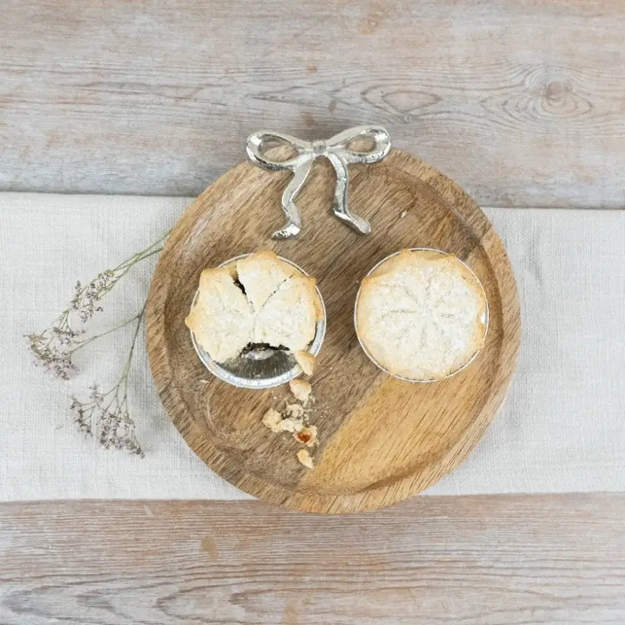 Two small baked pies in metal pie dishes on a round wooden serving board, with a silver bow-shaped spoon or utensil above them and dried flowers to the left, set on a light wood surface.