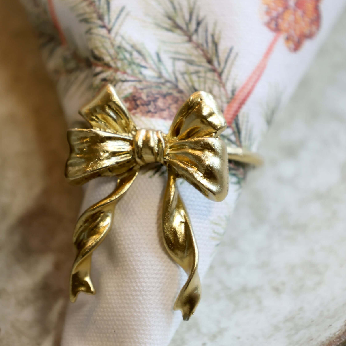 A close-up of a shiny gold bow-shaped ring holding a white napkin with a holiday pine branch and orange flower print, resting on a textured light surface.