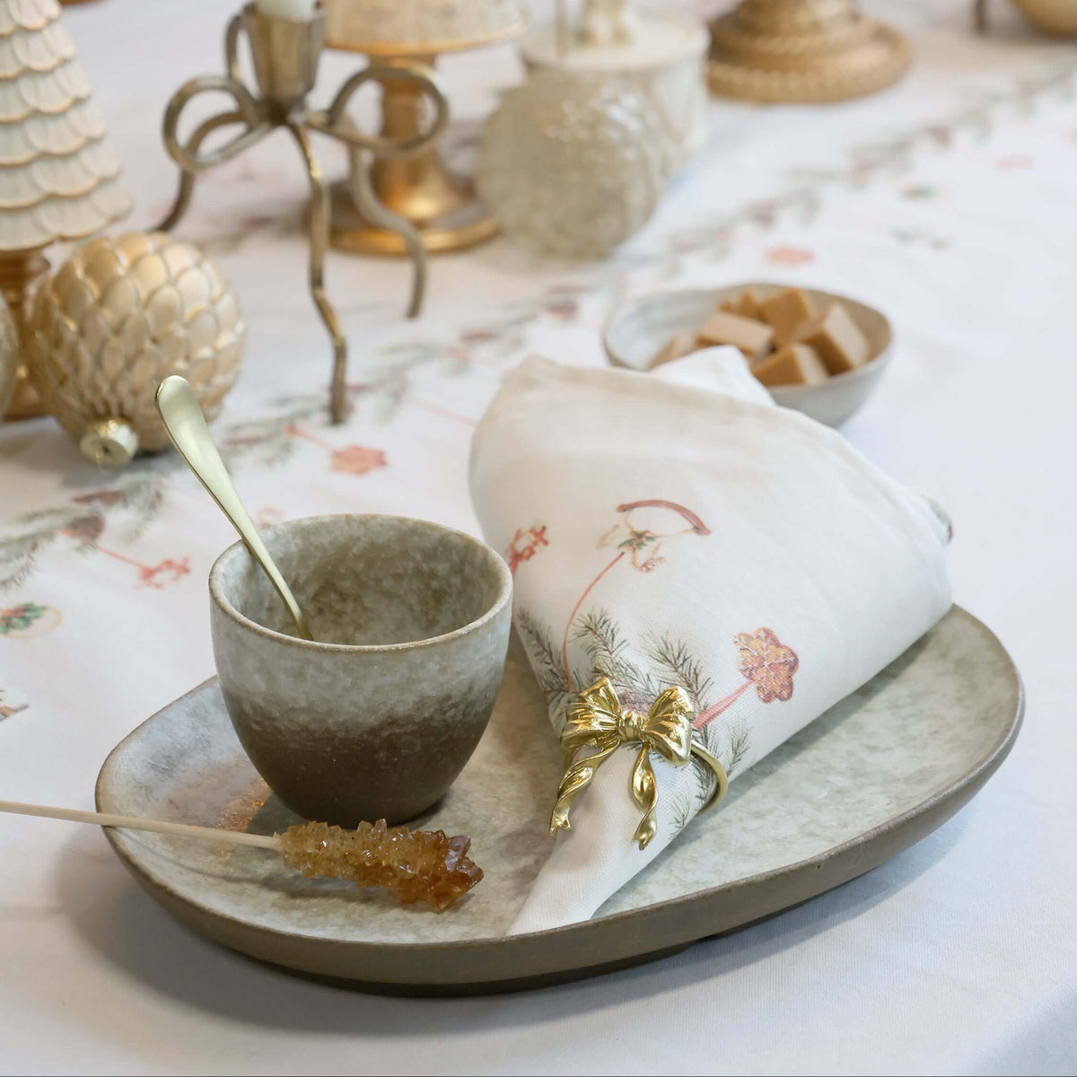 A styled holiday table setting featuring a ceramic plate with a speckled gray-green glaze, a matching napkin with delicate floral embroidery and a gold bow, a small speckled bowl, and a spoon. The table is decorated with cream textured Christmas tree ornaments, a nutcracker figurine, and warm candlelight.
