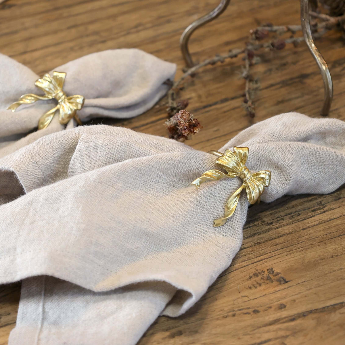 Two light beige linen napkins tied with decorative gold bow ring holders, displayed on a rustic wooden table.