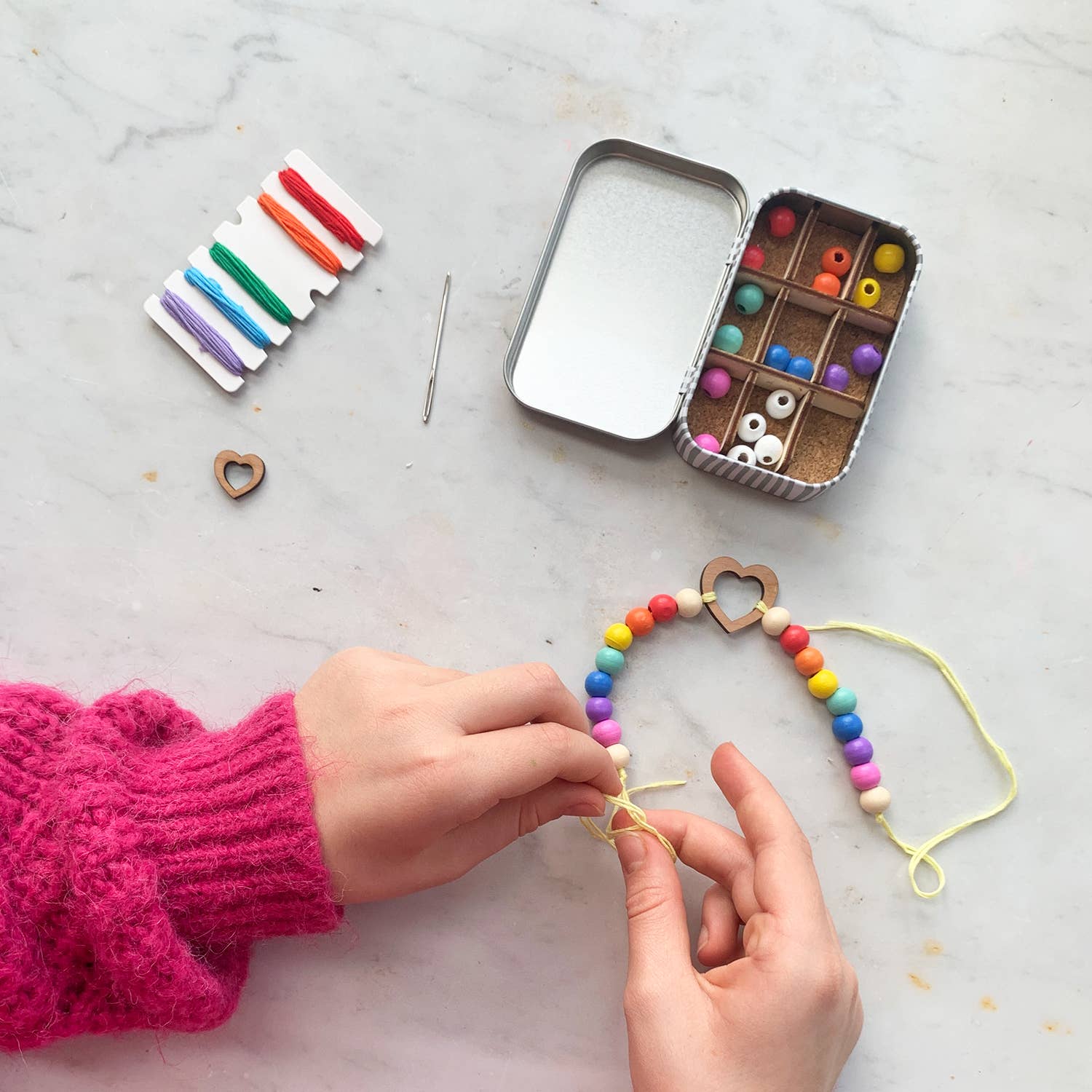 Person making a colorful bead necklace with a small tin of beads and a string of beads on a marble surface.
