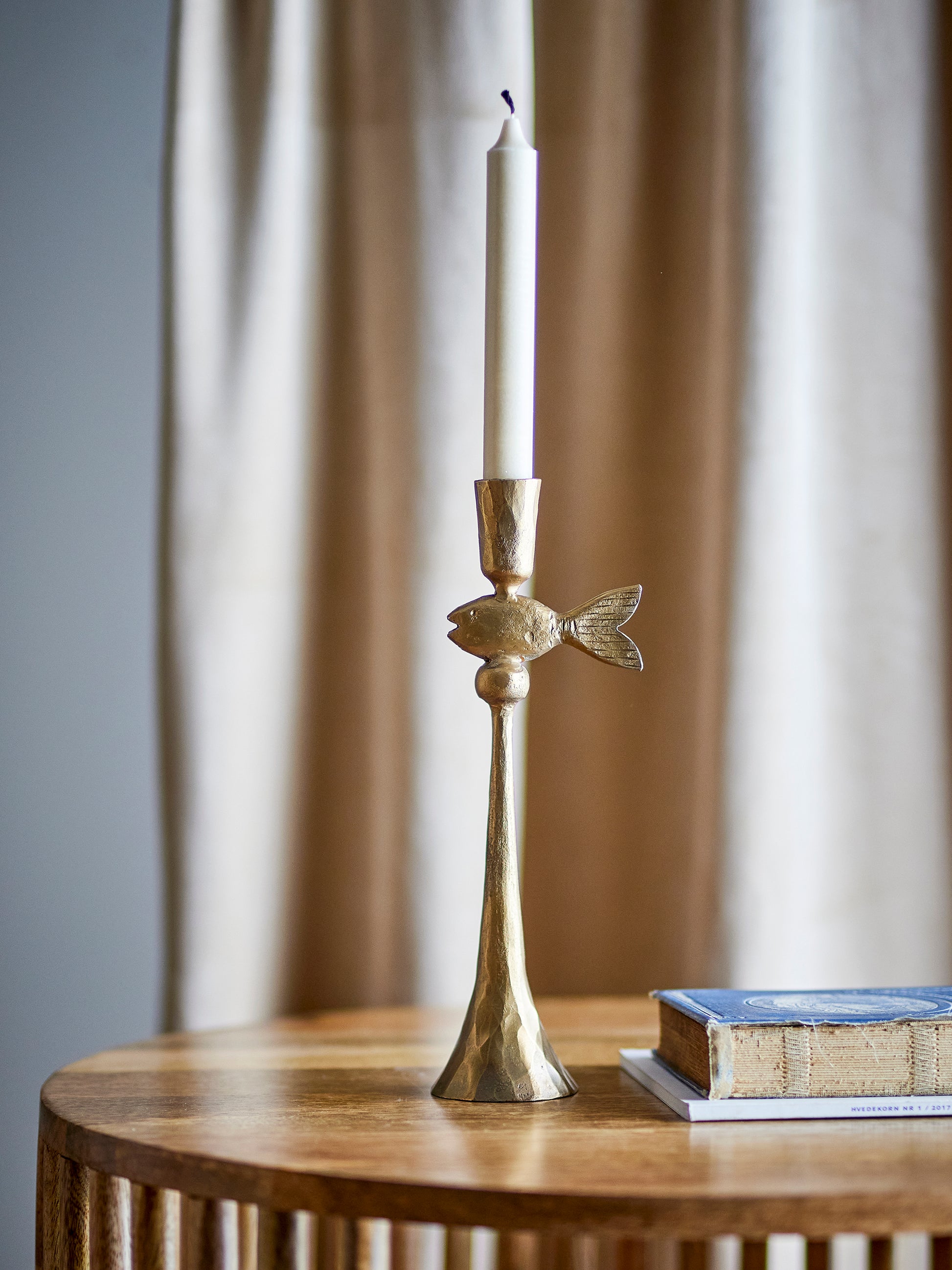 Brass candlestick holder with a white candle on a wooden surface with a blurred curtain background