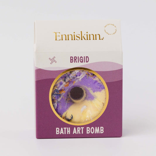 Enniskinn Brigid Bath Art Bomb in its original packaging, featuring a purple and cream-colored circular art bomb visible through a gold-trimmed window on a white and purple box.