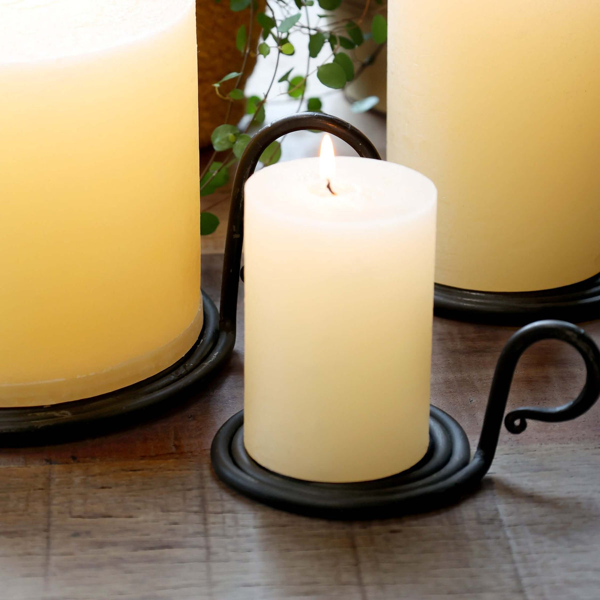 Three lit cream-colored pillar candles in black metal holders, arranged on a wooden surface with green trailing leaves in the background.