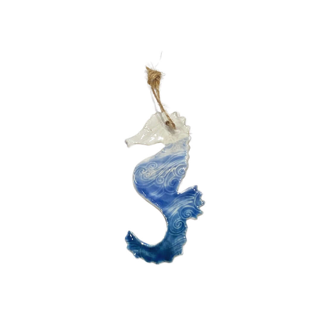 A ceramic seahorse decoration with an embossed wave pattern, in ombre blue and white colors, hanging from a jute string.
