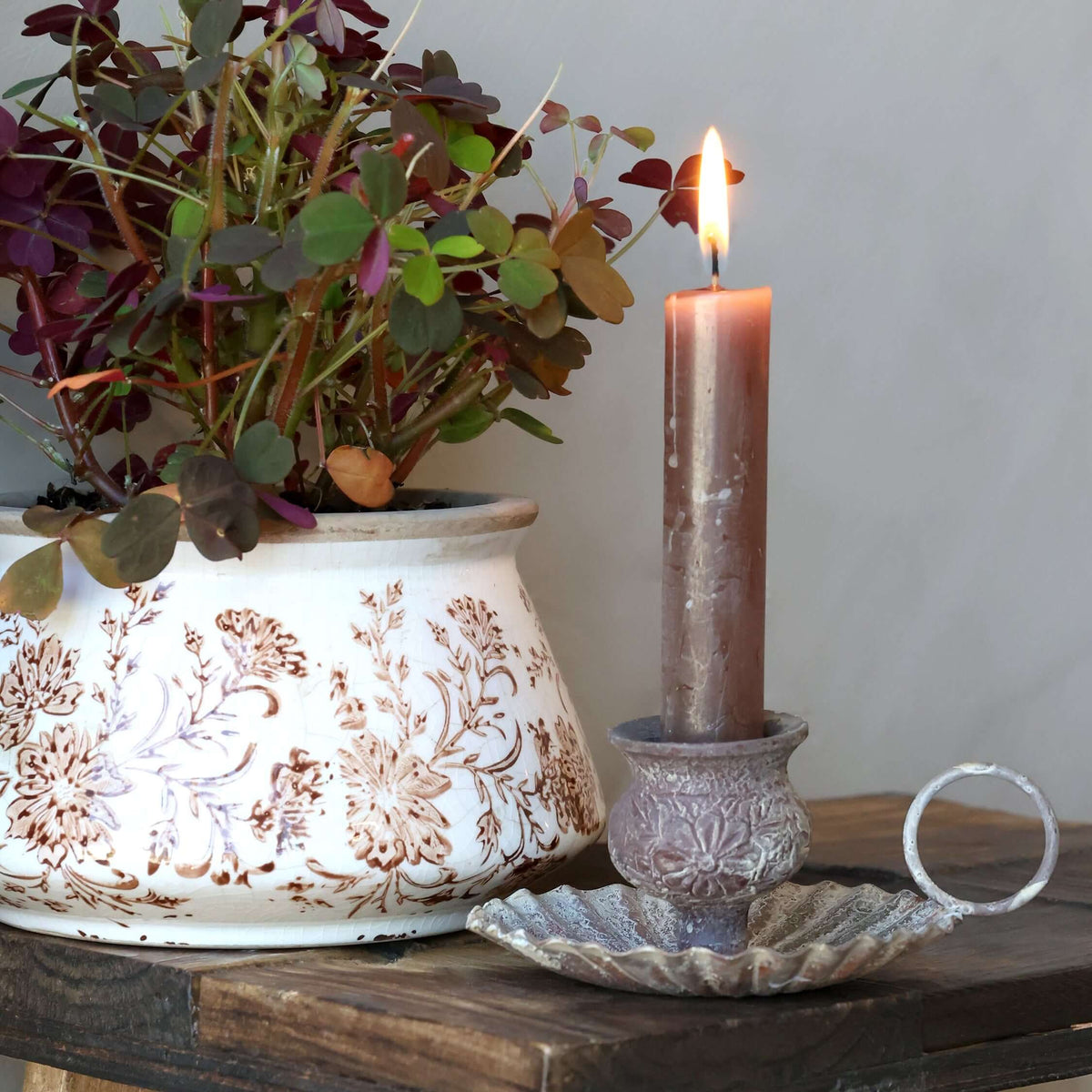 A lit brown pillar candle in a textured holder sits on a rustic wooden table next to a white ceramic planter with a brown floral pattern and a small silver ring, against a neutral gray wall.