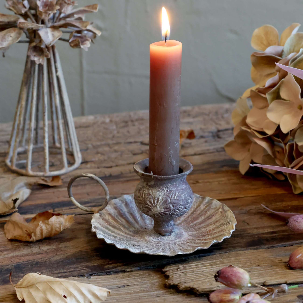 A lit taper candle in a textured, scalloped metal holder with a ring pull, styled on a rustic wooden table with dried flowers and leaves.