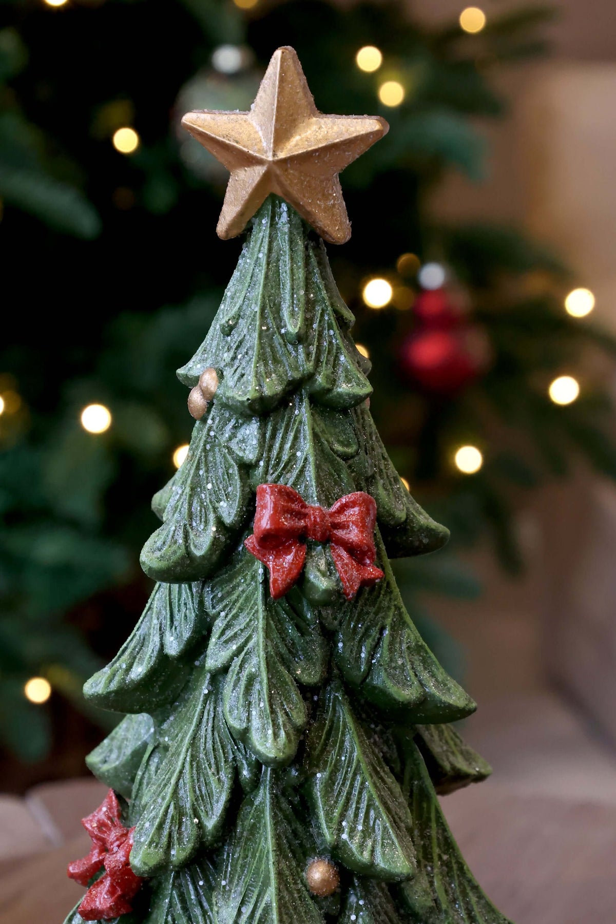 A green ceramic Christmas tree ornament with a textured leaf pattern, decorated with red bows, small gold ornaments, and topped with a gold star, shown against a blurred holiday background.