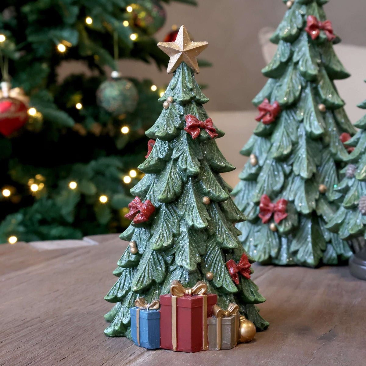 A green ceramic Christmas tree ornament with a gold star topper, decorated with red bows, gold beads, and small gift boxes at the base, displayed on a wooden surface with blurred Christmas trees in the background.
