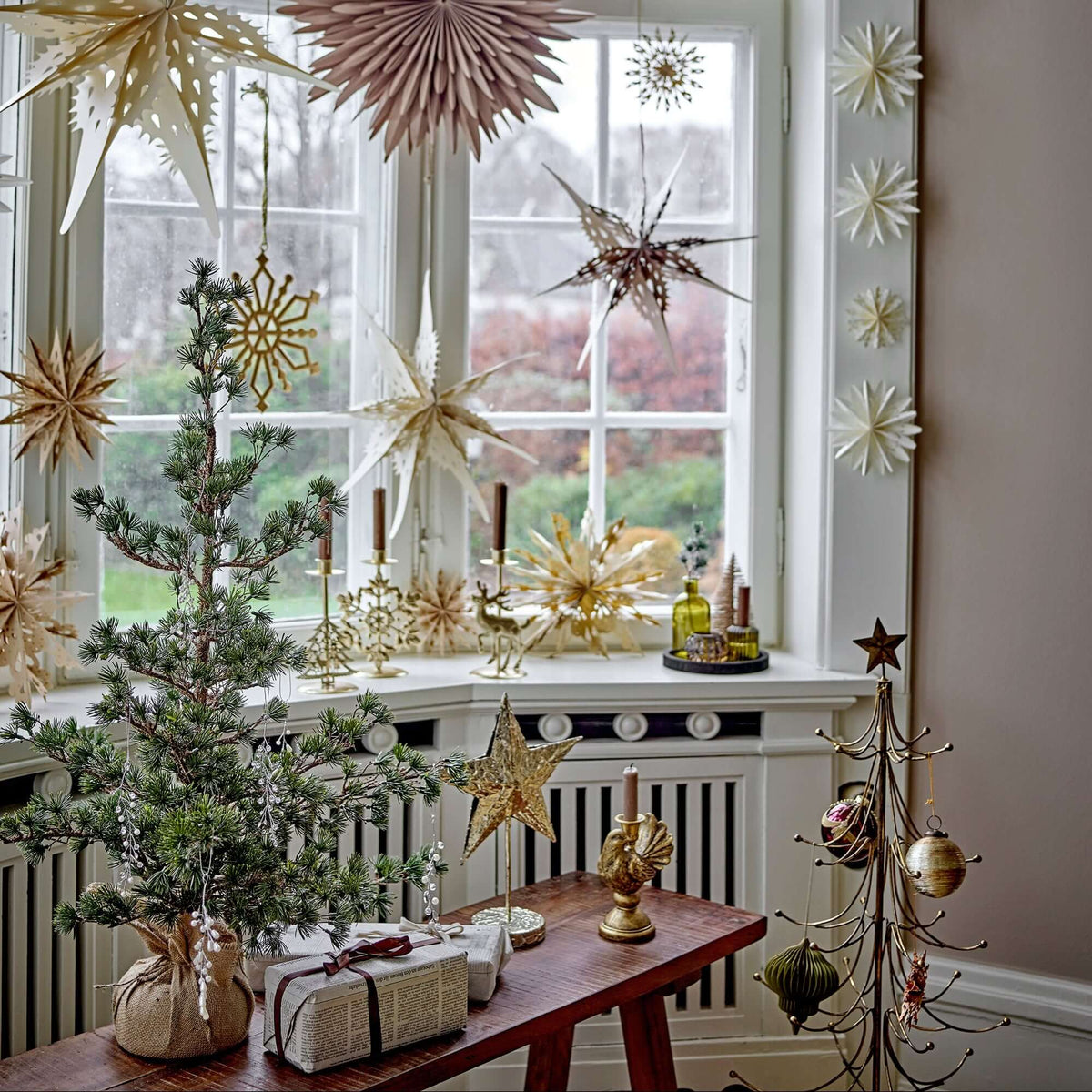 A styled holiday window display featuring gold and beige paper star ornaments hanging around a white-framed window, a small potted evergreen tree on a wooden table, wrapped gifts, candles, and decorative metal ornaments in a warm, festive interior setting.