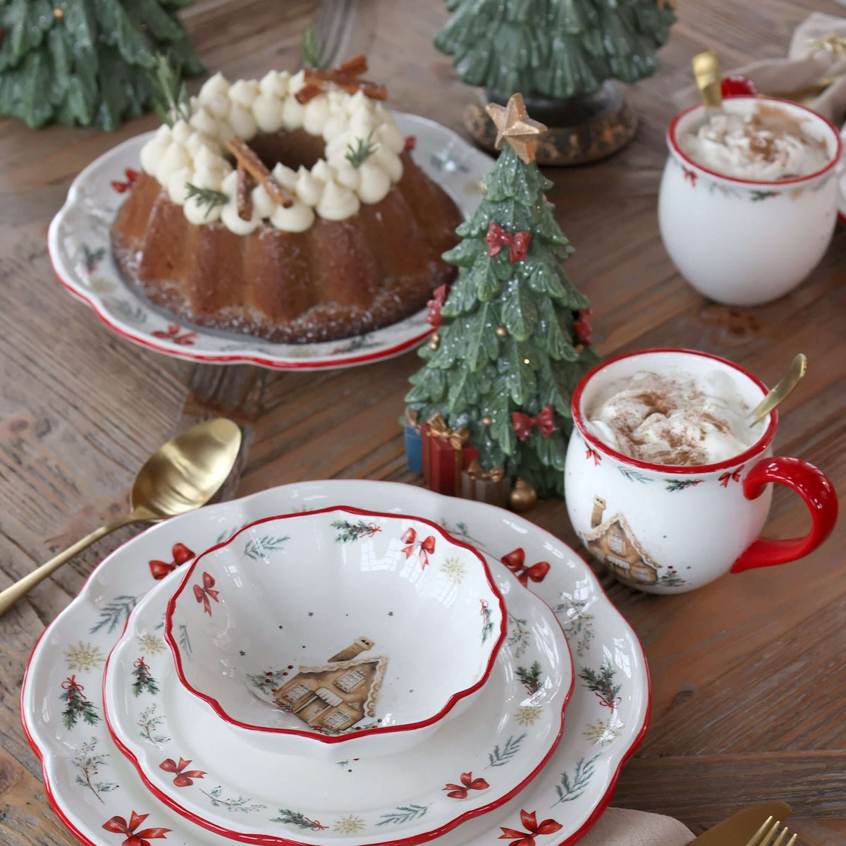A styled holiday table setting featuring a white ceramic dinnerware set with red rims, green pinecone motifs, and red berry accents, arranged on a rustic wooden table alongside gold-toned cutlery, mugs with whipped cream and cinnamon, and decorative Christmas trees.