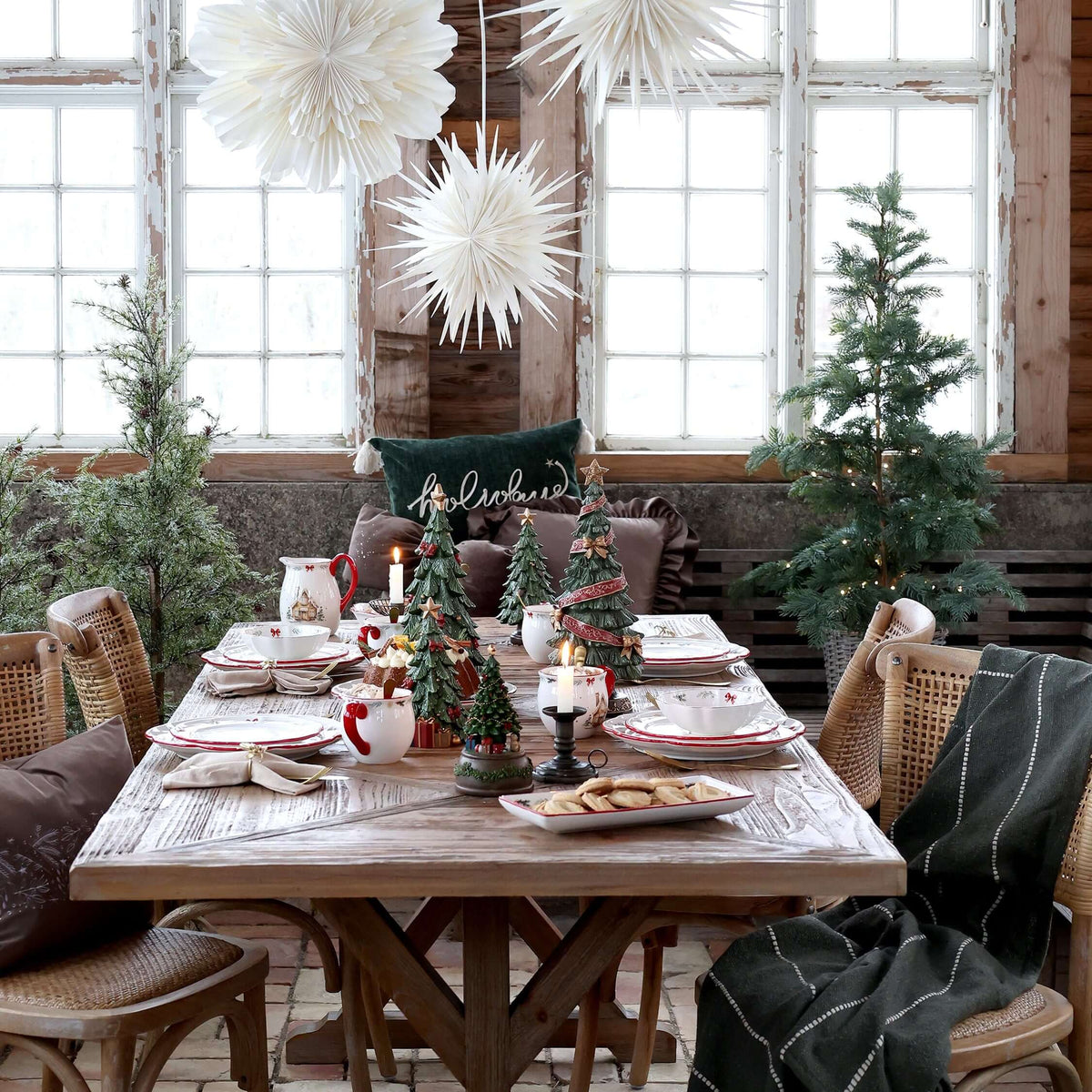 A rustic wooden dining table set for a holiday meal in a cozy room with large windows, hanging white paper lanterns, potted trees, and a dark green striped throw blanket draped over the chair.