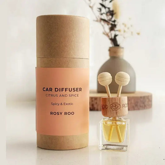 A car diffuser set featuring a cylindrical kraft paper packaging container with a peach label and a small glass diffuser bottle with wooden diffuser tops, shown on a light surface with a blurred vase in the background.