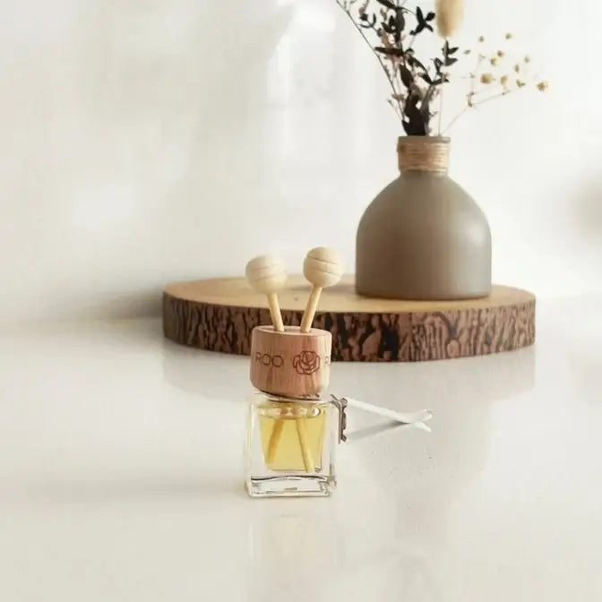 A small glass reed diffuser bottle with a light wood cap and two wooden diffuser sticks, placed on a white surface in front of a ceramic vase with dried stems on a round wood slice.