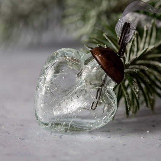 A clear crackle glass heart ornament with a bronze cap and organza tie, placed on a surface with a blurred greenery background.